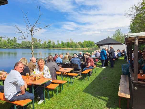 Gruppen von Menschen sitzen an Tischen am Wasser, umgeben von Bäumen und blauem Himmel.